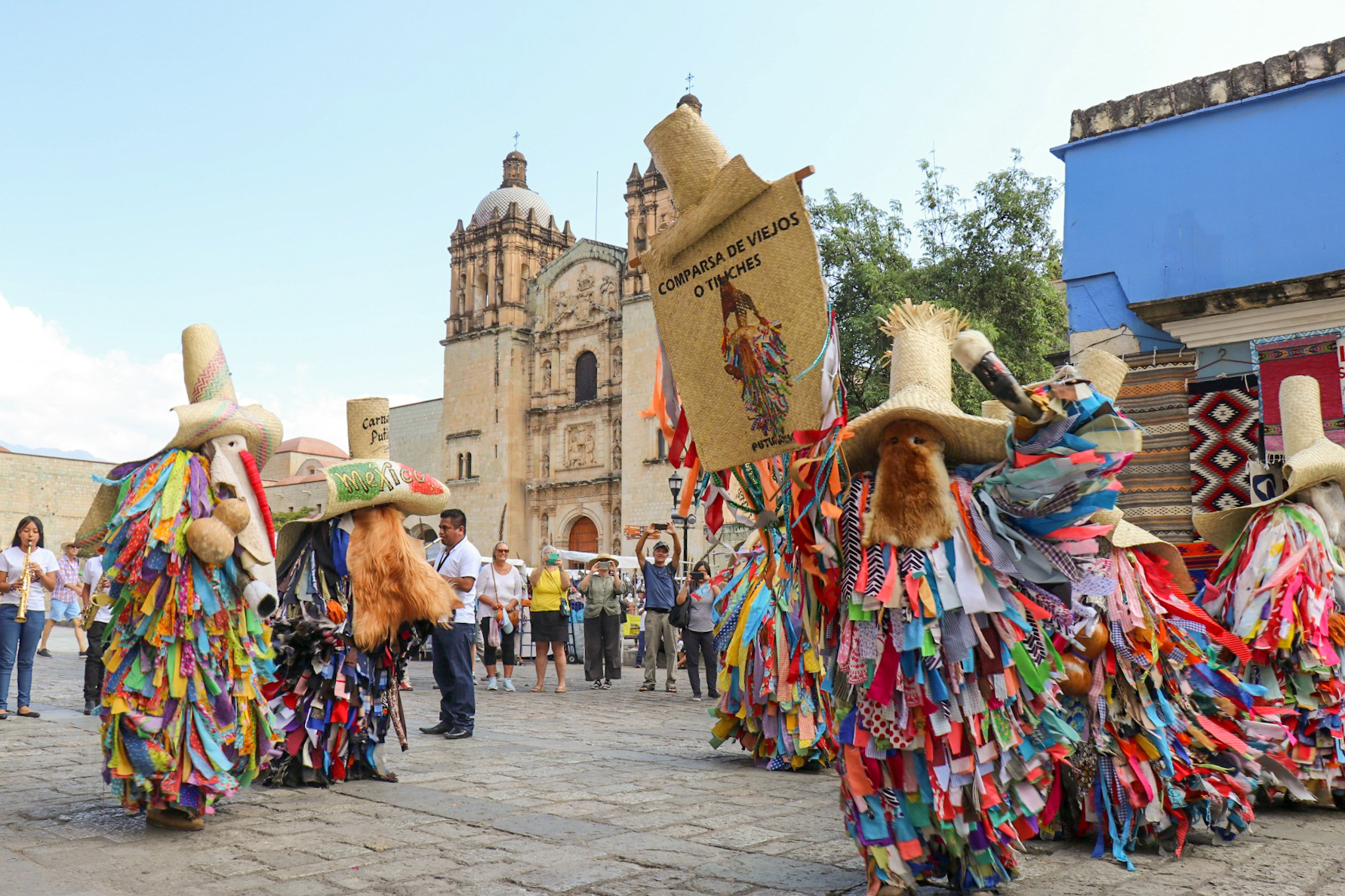 A masked Carnaval dancer in Oaxaca wearing cowbells while a brass band plays behind them on a village street