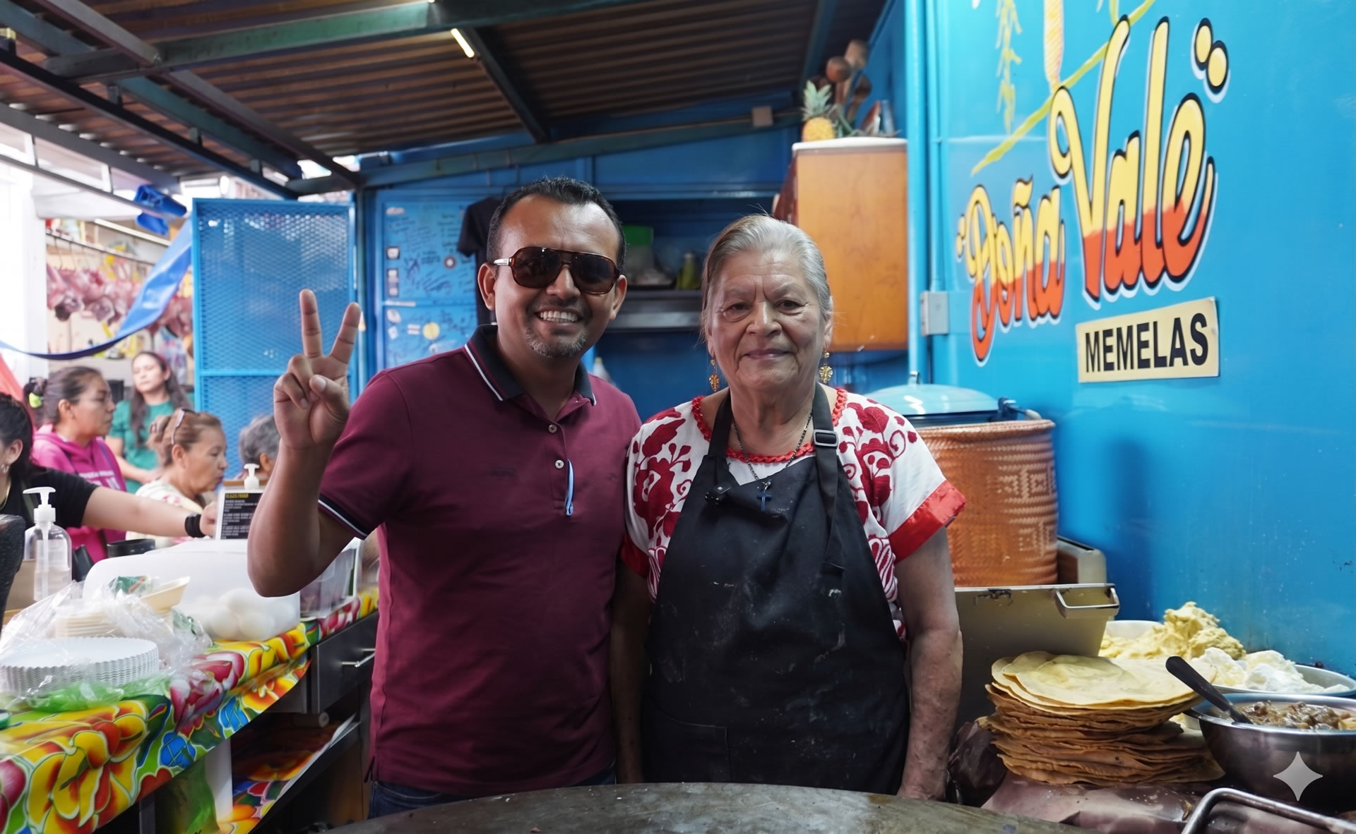 Doña Vale and her team preparing fresh memelas on the large comal at her stand inside Mercado Central de Abastos, with rows of sizzling tortillas and traditional toppings