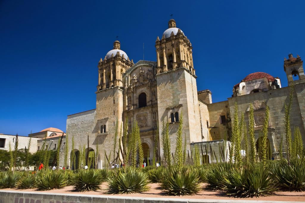 Pedestrian street view in Oaxaca historic center with Santo Domingo Church golden interior visible, nearby markets and colonial architecture under clear blue skies